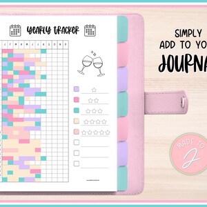 Könnte beinhalten: Ein rosa Planer mit einer Jahresübersichtsseite. Der Tracker hat ein Raster mit pastellfarbenen Blöcken und dem Text "YEARLY TRACKER". Der Planer enthält auch den Text "SIMPLY ADD TO YOUR JOURNAL".