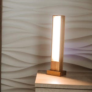 May include: A rectangular wooden bedside lamp with a bright, vertical light panel. The lamp is on a wooden nightstand next to a bed with a patterned pillow and a brown headboard. The wall behind the bed has a textured, wave-like design.