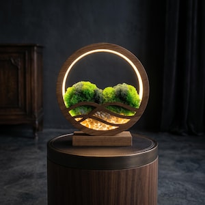 May include: A contemporary wooden lamp with a circular design. It has a glowing ring of light, illuminating green moss and small stones. The base is dark wood, and the lamp sits on a dark brown table. The lamp is a decorative item.