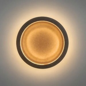 Op de afbeelding: Een ronde wandlamp met een warme, gouden gloed. De lamp heeft een donker, cirkelvormig frame met een lichter, getextureerd interieur. Het is gemonteerd op een grijze muur, wat een moderne en elegante uitstraling creëert.