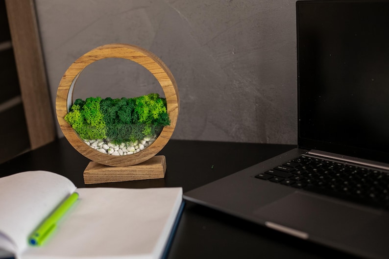 May include: A wooden, circular decorative piece with a natural moss and stone arrangement. The moss is various shades of green, and the stones are white. The piece is on a wooden base. A laptop and notebook with a pen are also on the desk.