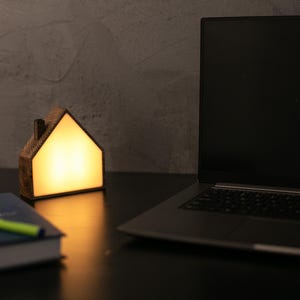 May include: A wooden house-shaped lamp with a warm, glowing interior sits on a dark surface next to a laptop and a book with a green pen. The lamp's design resembles a small house.