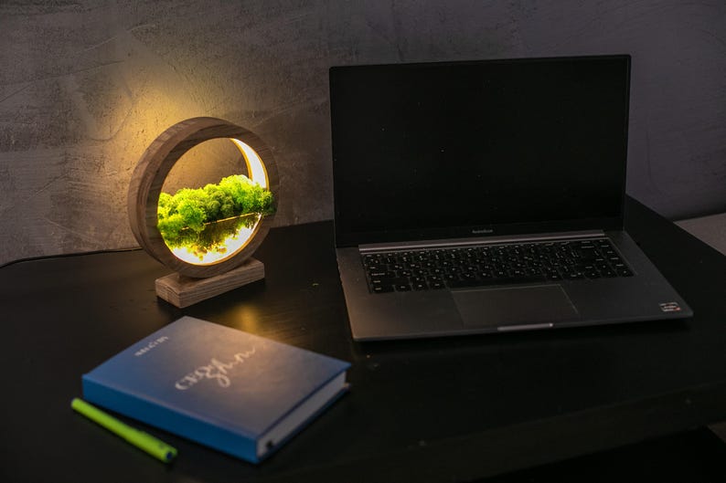 May include: A wooden desk lamp with a circular design, featuring a bright light illuminating green moss. A laptop, a blue notebook, and a green pen are on the black desk. The lamp has a natural, earthy aesthetic.