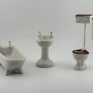 May include: A miniature bathroom set featuring a bathtub, sink, and toilet. The white porcelain pieces have gold-colored accents on the faucets and toilet chain. The bathtub has four small feet. The toilet has a brown rim.