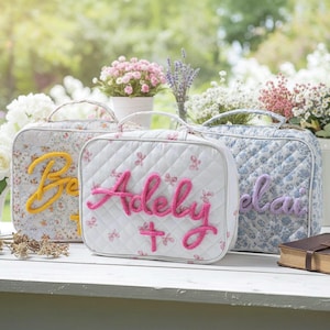 May include: Three floral patterned cosmetic bags with embroidered names. The center bag is white with pink embroidery, the left bag is floral with yellow embroidery, and the right bag is blue floral with purple embroidery. The bags have handles and are on a white surface.