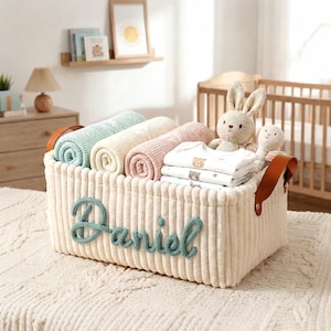 Puede incluir: Una cesta de almacenamiento de pana color crema con el nombre "Daniel" en escritura turquesa. La cesta contiene mantas enrolladas en colores pastel, ropa de bebé y un conejito de peluche. Asas de cuero marrón.