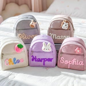 May include: Five corduroy backpacks in various colors, each with a name embroidered on the front. The backpacks feature decorative patches of a strawberry, flower, bunny, giraffe, and hedgehog. The names are Oliver, Chloe, Mason, Harper, and Sophia.