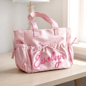 May include: A pale pink satin tote bag with a top handle and side pockets. The name "Evelyn" is embroidered in a darker pink script on the front. The bag is on a light wood surface.