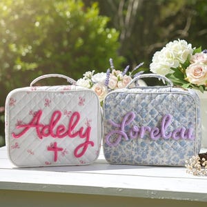 May include: Two quilted cosmetic bags with handles sit on a white table. One is white with pink floral accents and the name "Adely" in pink script. The other is blue with a floral pattern and the name "Lorelay" in purple script.