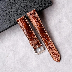 Puede incluir: Dos correas de reloj de cuero marrón con un patrón texturizado, con una hebilla plateada. Las correas están colocadas sobre una superficie gris claro junto a una caja negra. Las correas están hechas de material similar a reptiles.