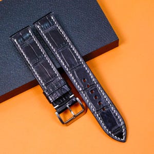 May include: Two black watch straps with white stitching and a silver buckle. The straps have a textured, crocodile-like pattern. One strap is fastened, and the other is unfastened. The background is orange and dark gray.