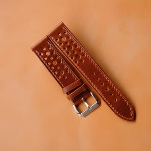 Könnte beinhalten: Ein beiges Leder-Uhrenarmband mit perforiertem Design und mehreren kreisförmigen Ausstanzungen entlang des Bandes. Das Armband verfügt über zwei Lederschlaufen und eine polierte silberfarbene Metallschnalle zur sicheren Befestigung.