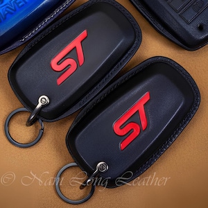 May include: Two black leather key fob covers featuring a prominent red ST logo embossed on the front. Each cover includes a dark metal ring attachment secured by a small screw. The design is sleek with visible stitching along the edges.