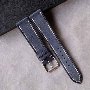 Può includere: Un cinturino blu navy con cuciture bianche e fibbia argentata. Il cinturino è realizzato in un materiale strutturato e viene esposto su una superficie grigia. Il cinturino è nuovo e inutilizzato.