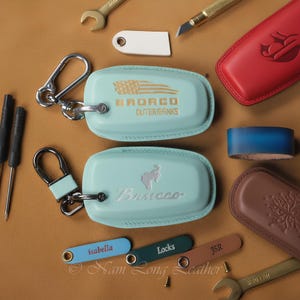 May include: Two light blue leather key fob covers with metal clips. One cover features a debossed American flag and the text BRONCO OUTERBANKS. The other cover displays a bucking horse logo and the word Bronco. Also included are small leather tags with names.
