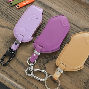 May include: Three leather car key fob covers in light purple, dark purple, and tan. Each cover features a matching leather strap attached to a metal clasp or ring. The covers have embossed logos and are displayed alongside small colourful key tags.
