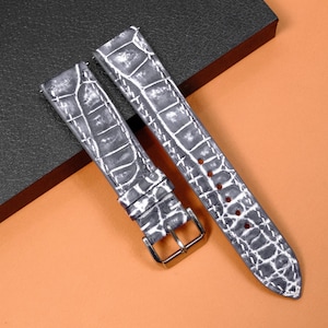 Puede incluir: Una correa de reloj de dos piezas con un patrón de relieve de caimán en gris y blanco. La correa incluye una hebilla de metal plateado, una trabilla deslizante a juego y costuras blancas en contraste a lo largo de los bordes.
