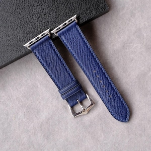 May include: A pair of deep blue leather watch straps with silver-colored metal clasps and connectors. The straps have a textured surface and are displayed on a light gray surface next to a dark gray box.