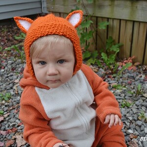 May include: A baby wearing an orange fox costume with a knitted orange hat with white ears. The baby is sitting on a bed of gravel.