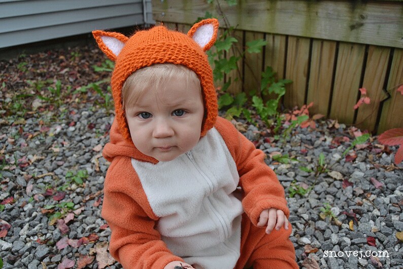 May include: A baby wearing an orange fox costume with a knitted orange hat with white ears. The baby is sitting on a bed of gravel.
