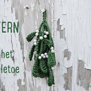 May include: A green crocheted mistletoe sprig with white pearl beads hanging from a white wooden surface. The text "PATTERN Crochet Mistletoe" is visible in the image.