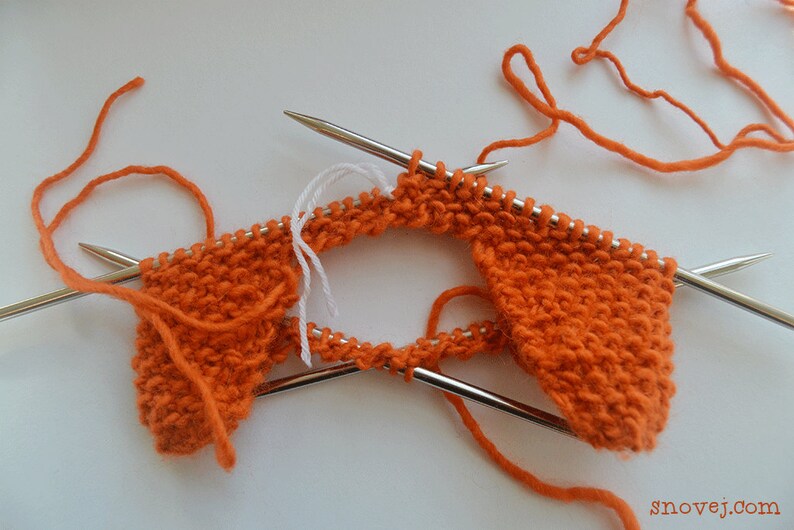 May include: Orange knitting project in progress with two knitting needles and white yarn. The knitting is in a circular shape with two ends.
