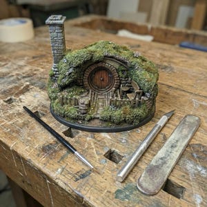 Può includere: Un modello in miniatura di una casa hobbit con porta rotonda, camino in pietra e tetto muschioso. Un pennello, un taglierino e una lima metallica sono su un banco da lavoro in legno. Tema fantasy.