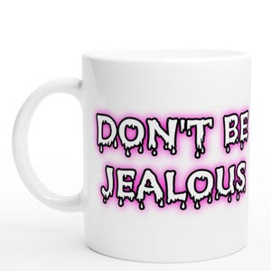 May include: White ceramic mug featuring the phrase "DON'T BE JEALOUS" in a dripping font with a pink neon outline. The mug has a curved handle and a glossy finish. The text is in black with a pink glow.