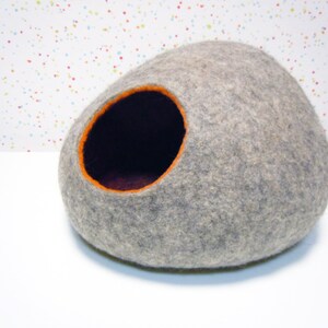 May include: A gray felt cat bed shaped like a rounded stone with an orange felt opening. The bed is on a white surface.