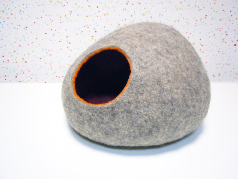 May include: A gray felt cat bed shaped like a rounded stone with an orange felt opening. The bed is on a white surface.