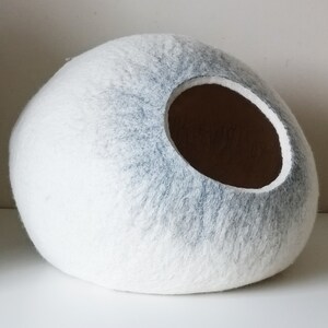 May include: A white and gray felt cat bed shaped like an egg with a round opening. The bed is made of felted wool and has a soft, fuzzy texture.