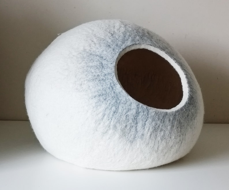 May include: A white and gray felt cat bed shaped like an egg with a round opening. The bed is made of felted wool and has a soft, fuzzy texture.