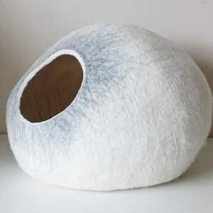 May include: A white and gray felt cat bed shaped like a large, round stone. The bed has a large opening on one side for the cat to enter.