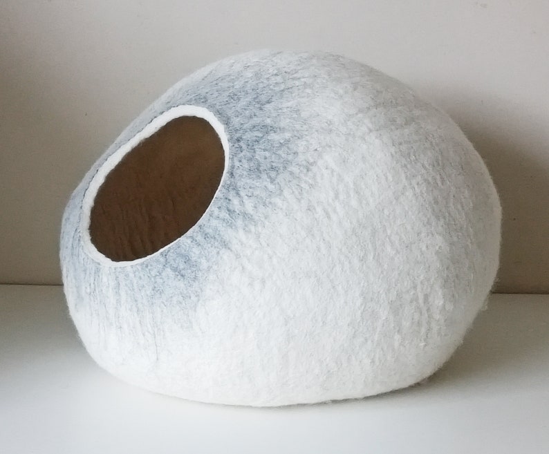 May include: A white and gray felt cat bed shaped like a large, round stone. The bed has a large opening on one side for the cat to enter.