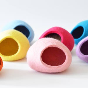 Puede incluir: Un conjunto de camas para mascotas de fieltro de colores, incluyendo naranja, amarillo, rosa, azul y morado. Las camas tienen forma ovalada con una abertura redonda en el centro.