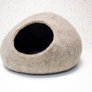 Pode incluir: Uma cama para gato de feltro bege em forma de oval arredondado com uma abertura de feltro preto. A cama est&aacute; sobre uma superf&iacute;cie branca com um fundo salpicado de cores.
