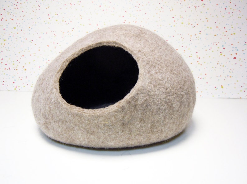 Pode incluir: Uma cama para gato de feltro bege em forma de oval arredondado com uma abertura de feltro preto. A cama est&aacute; sobre uma superf&iacute;cie branca com um fundo salpicado de cores.