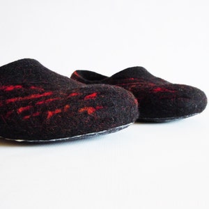 May include: A pair of black felt slippers with red accents. The slippers have a rounded toe and a slip-on design. The red detailing is in a linear pattern across the top of the slipper. The slippers are on a white background.