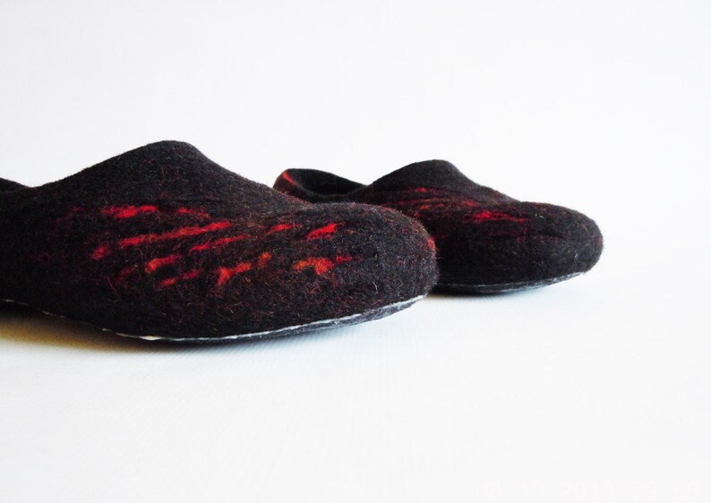 May include: A pair of black felt slippers with red accents. The slippers have a rounded toe and a slip-on design. The red detailing is in a linear pattern across the top of the slipper. The slippers are on a white background.