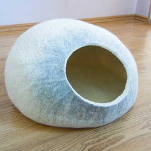 May include: A white and gray felt cat bed shaped like a dome with a large opening. The bed is on a wooden floor.