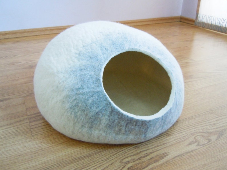 May include: A white and gray felt cat bed shaped like a dome with a large opening. The bed is on a wooden floor.