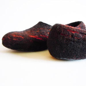 May include: A pair of felted slippers in a dark brown color with vibrant red accents. The slippers have a rounded toe and a low profile, suggesting a comfortable, indoor design. The texture appears soft and warm.