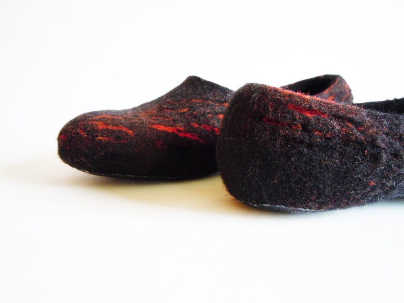 May include: A pair of felted slippers in a dark brown color with vibrant red accents. The slippers have a rounded toe and a low profile, suggesting a comfortable, indoor design. The texture appears soft and warm.