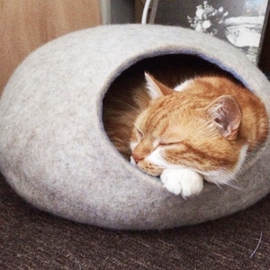Pode incluir: Uma cama para gatos de feltro cinza claro em forma de c&uacute;pula com uma pequena abertura. Um gato ruivo com patas brancas est&aacute; dormindo dentro da cama.