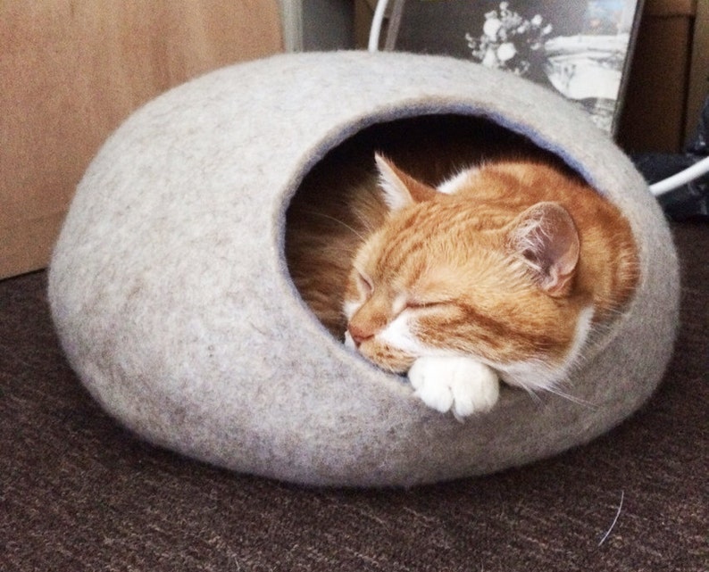Pode incluir: Uma cama para gatos de feltro cinza claro em forma de c&uacute;pula com uma pequena abertura. Um gato ruivo com patas brancas est&aacute; dormindo dentro da cama.