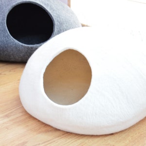 May include: A white felt cat bed shaped like a rounded oval with a circular opening. The bed is on a wooden floor.