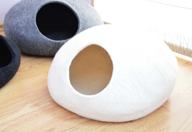 May include: A white felt cat bed shaped like a rounded oval with a circular opening. The bed is on a wooden floor.