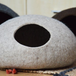 May include: A light gray felt cat bed with a round opening. The bed is on a woven rug with a dark blue border.