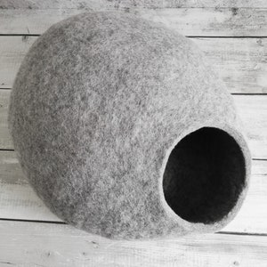 May include: A gray felt cat bed shaped like a cave with a round opening. The bed is on a white wooden surface.