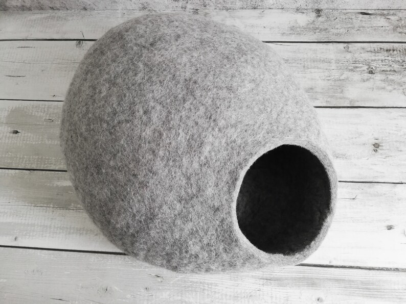 May include: A gray felt cat bed shaped like a cave with a round opening. The bed is on a white wooden surface.